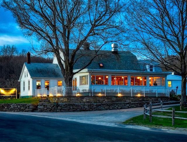 Lit white farmhouse with warm orange windows at dusk under bare trees