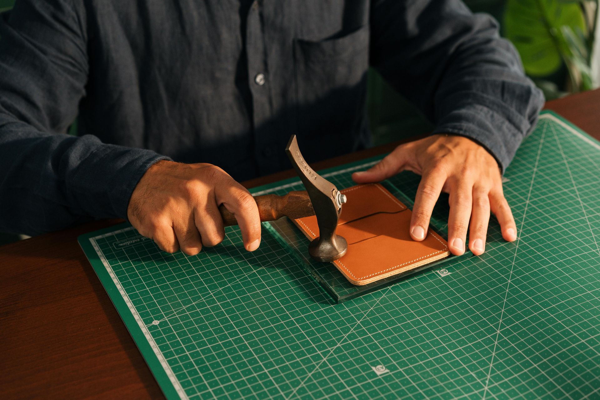 Hands using a hammer on a leather piece over a green cutting mat, crafting on a worktable.