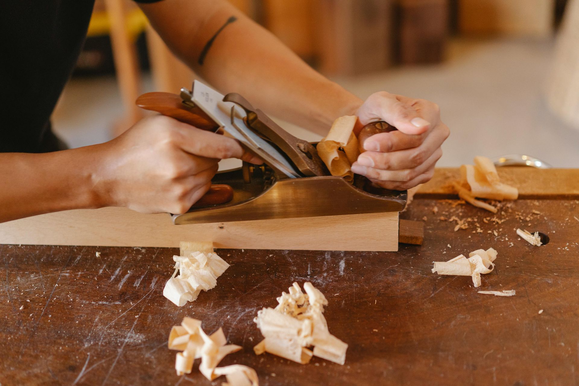 Hands planing a piece of wood on a workbench, with wood shavings scattered around.