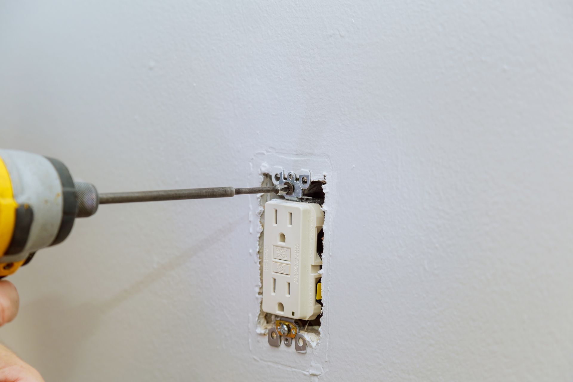 A hand uses a drill to remove a wall outlet from a white wall.