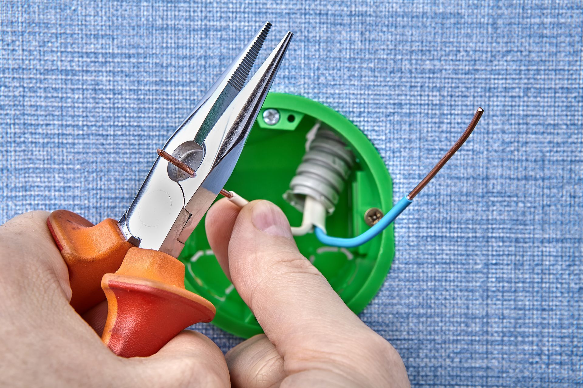 Hands holding pliers and a screwdriver inside a green electrical outlet box with exposed wires