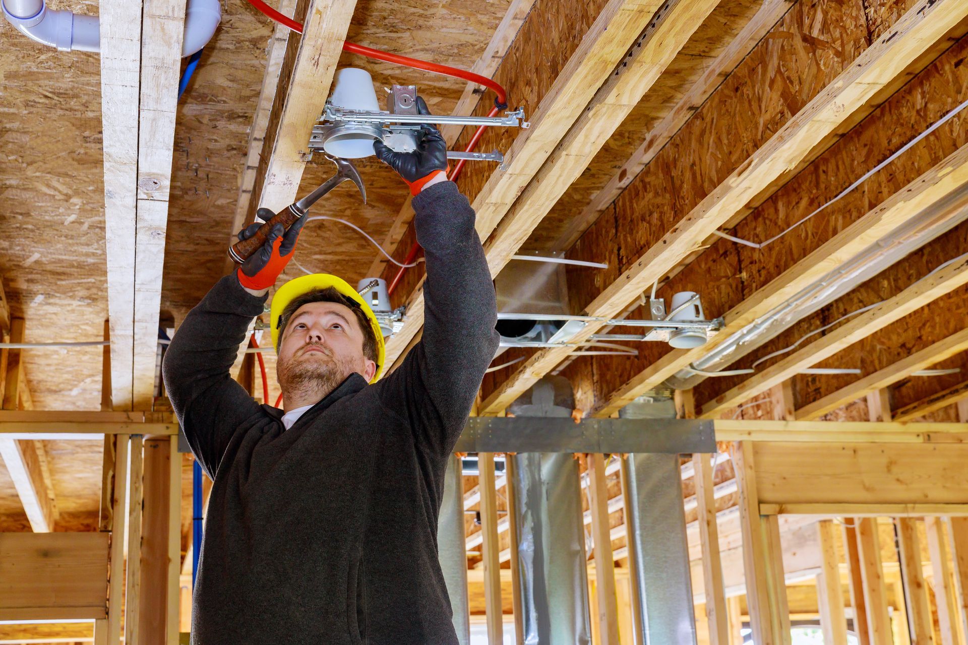 Construction worker installing wiring in a wooden house frame, wearing a hard hat and gloves.