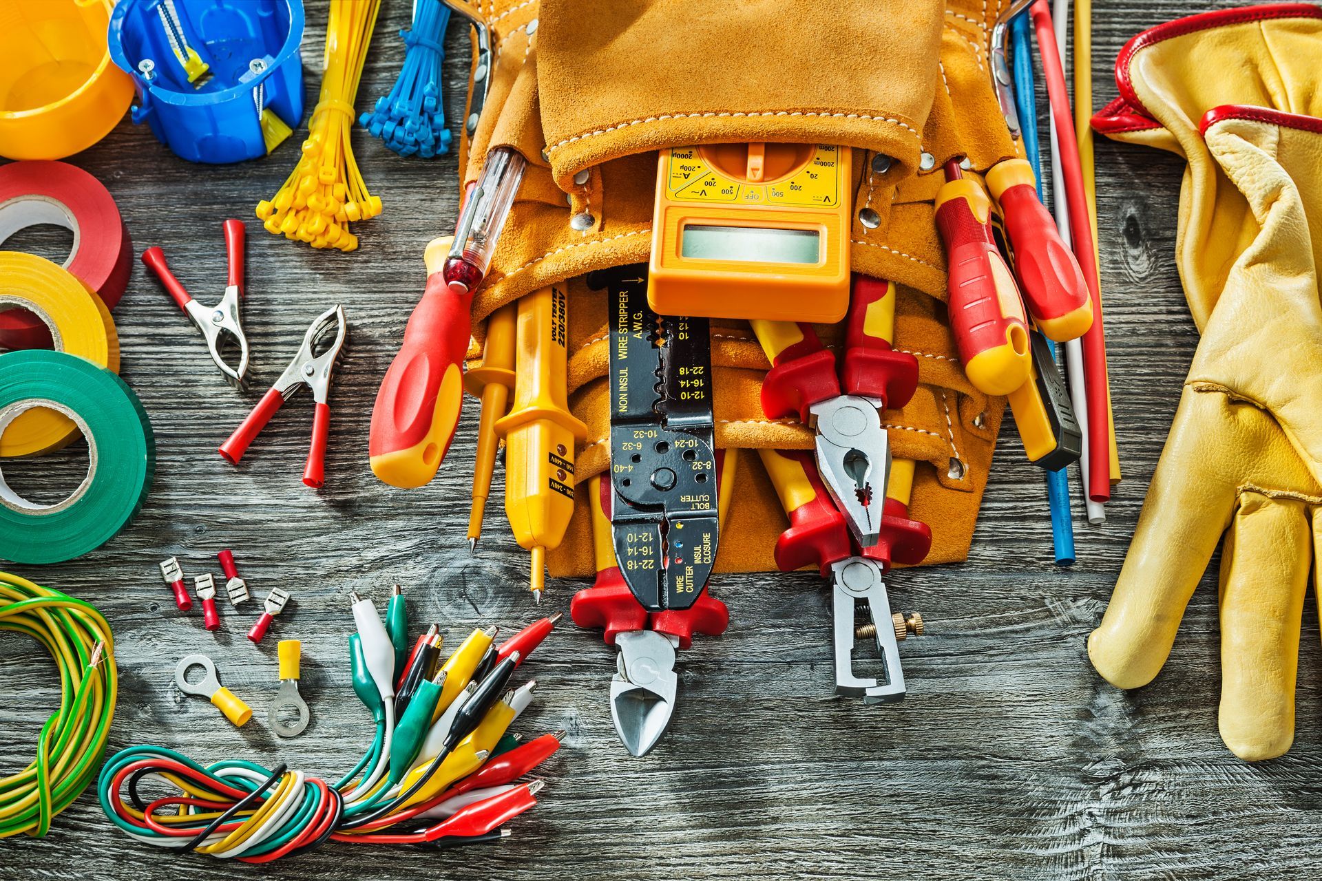 Assorted colorful tools and gloves arranged on a wooden surface, including pliers, screwdrivers, tape, and wires