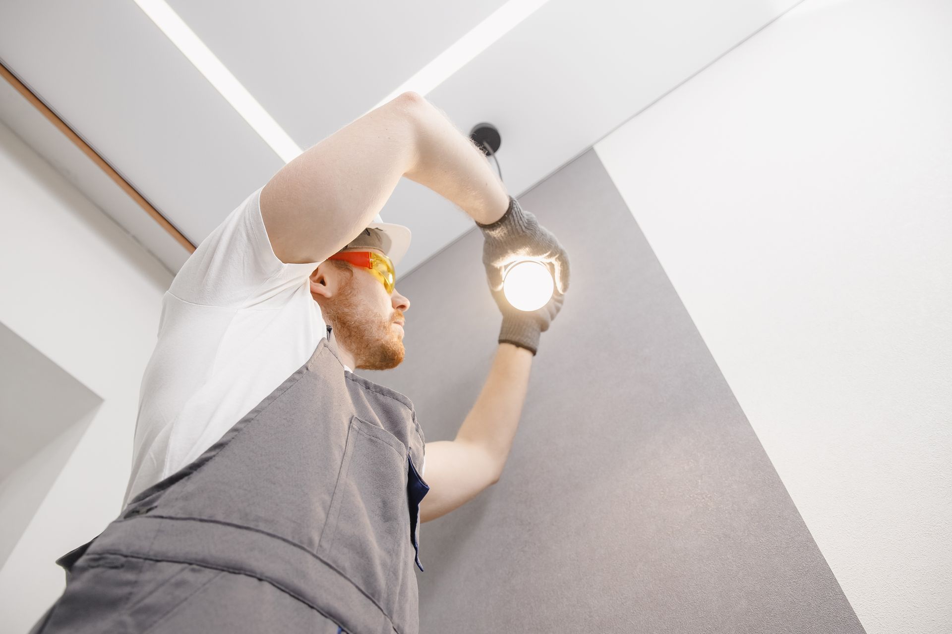 Worker in gray shirt and orange gloves installing a ceiling light indoors