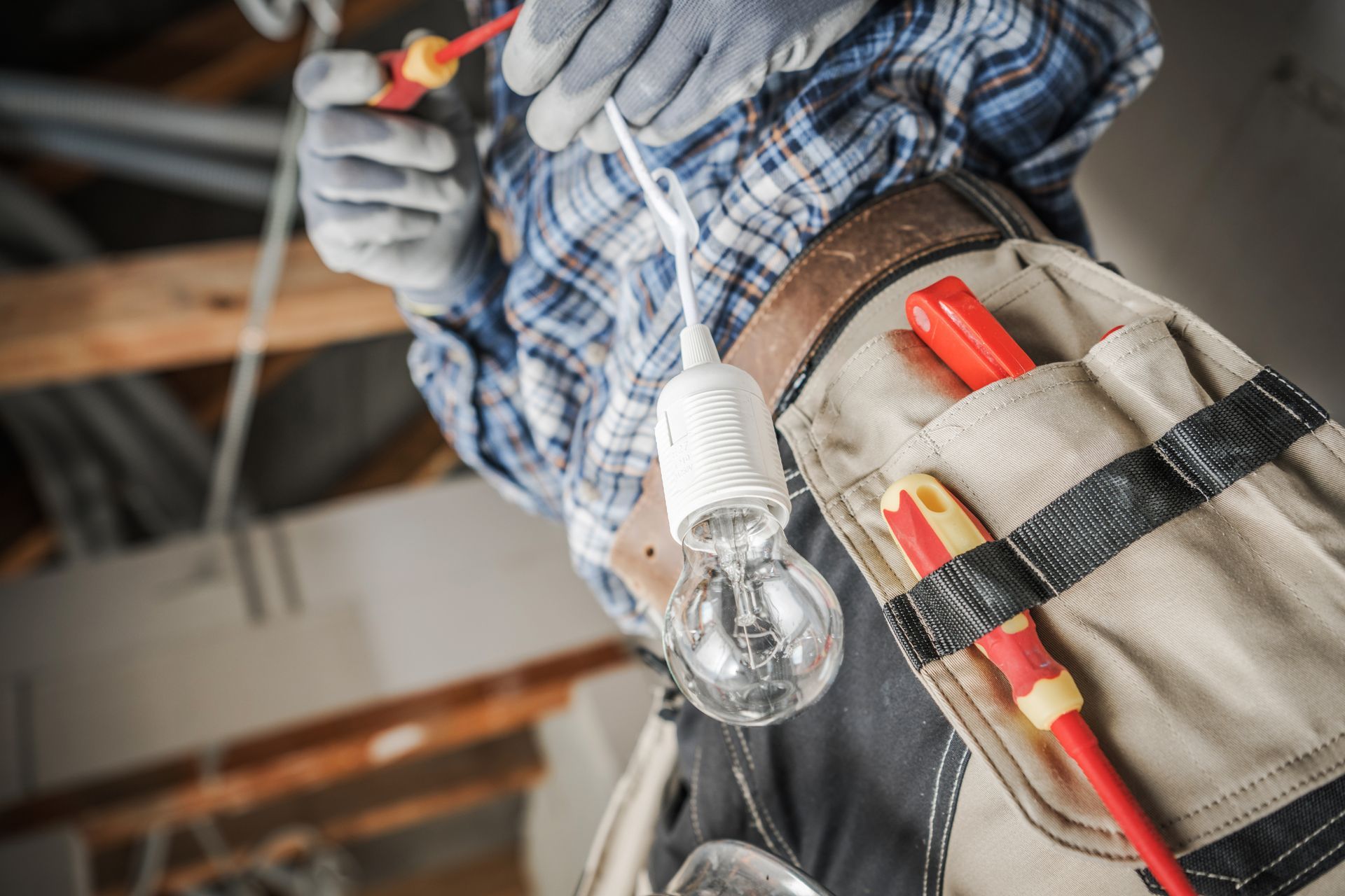 Electrician installing a light bulb while holding wires, wearing gloves and a tool belt with red tools