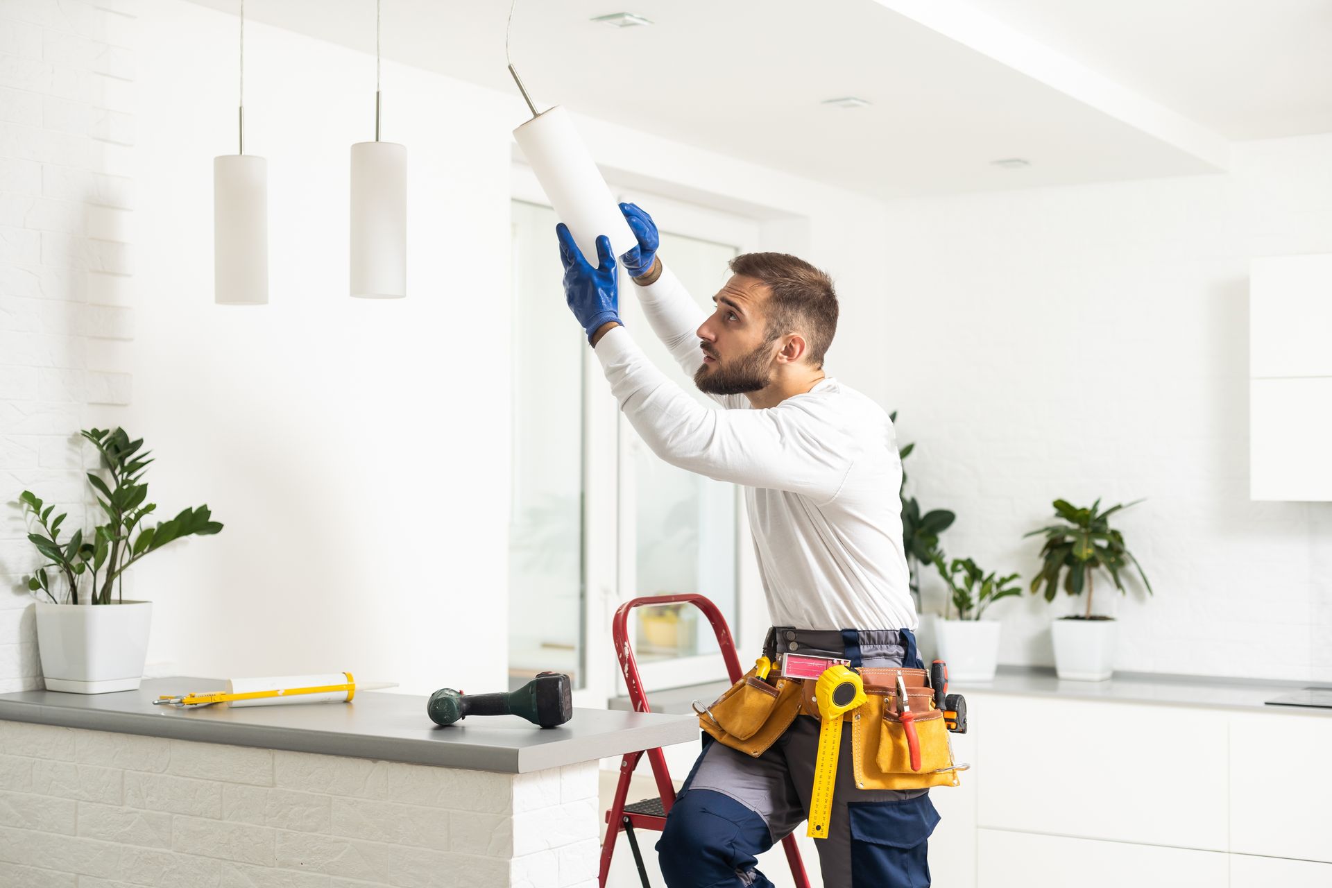 Electrician installing a ceiling light in a bright kitchen, holding a drill and wearing a tool belt