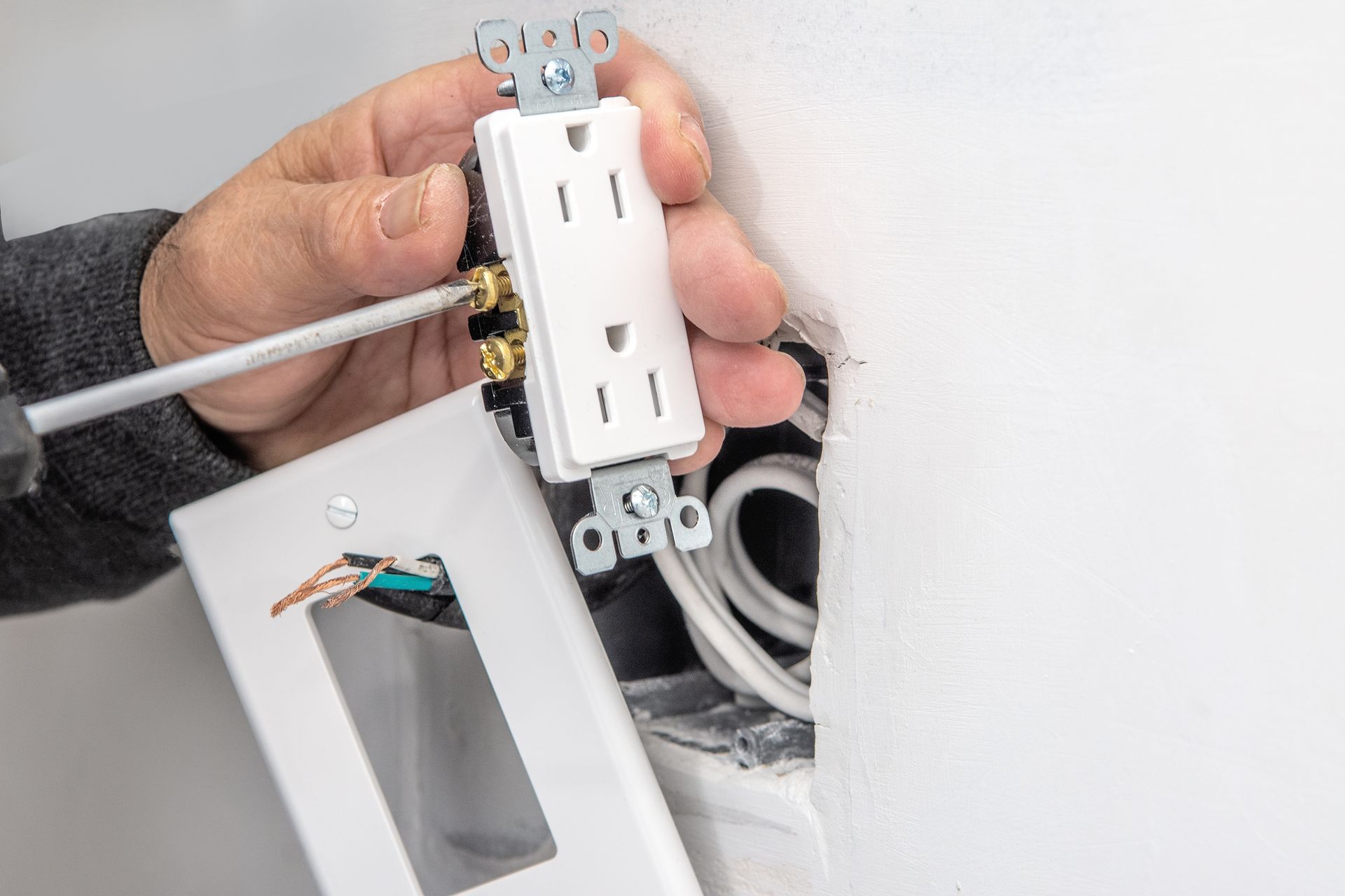 Hand holding a white electrical outlet with exposed wires near a wall opening