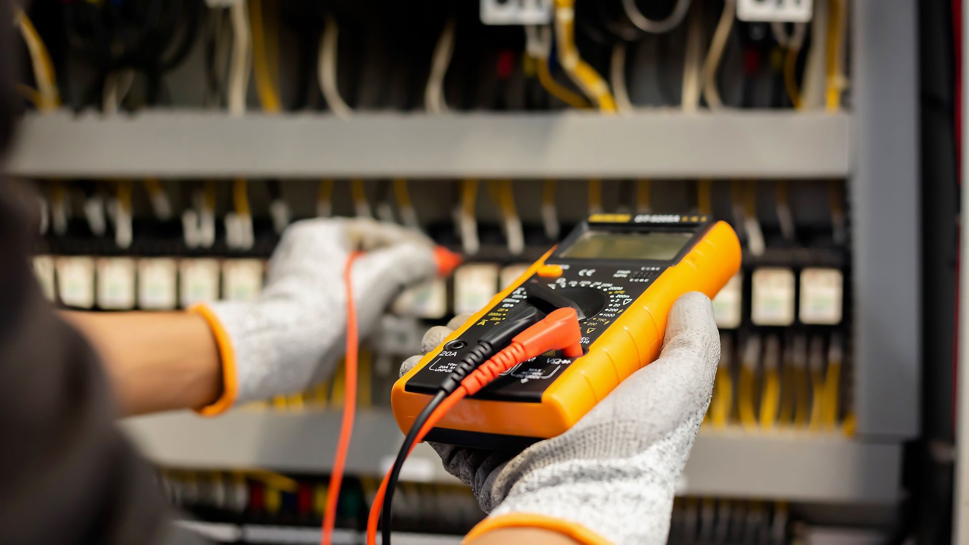 Gloved hands using a multimeter to test electrical wires in a control panel