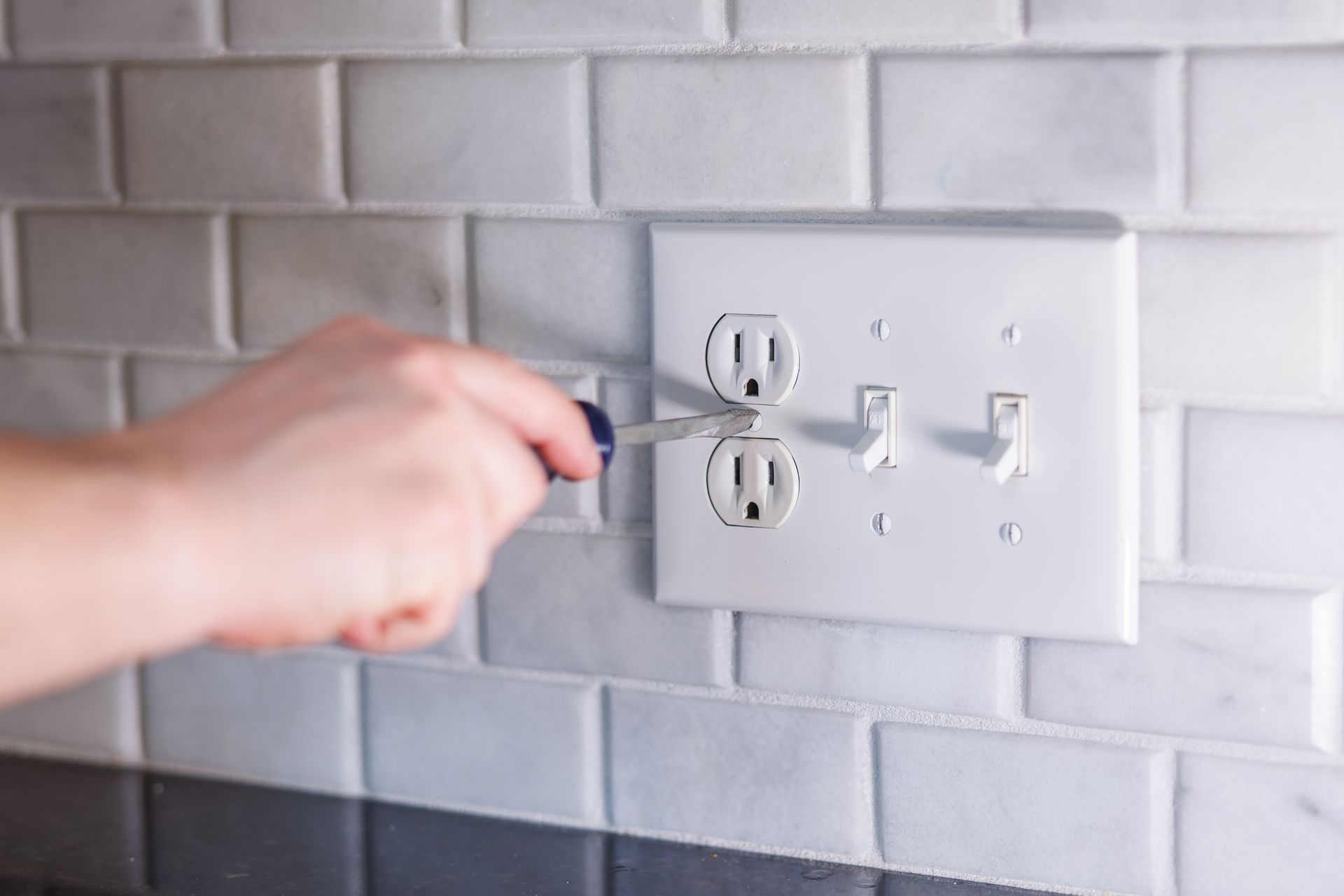Hand plugging a cord into a white wall outlet on a gray tiled backsplash