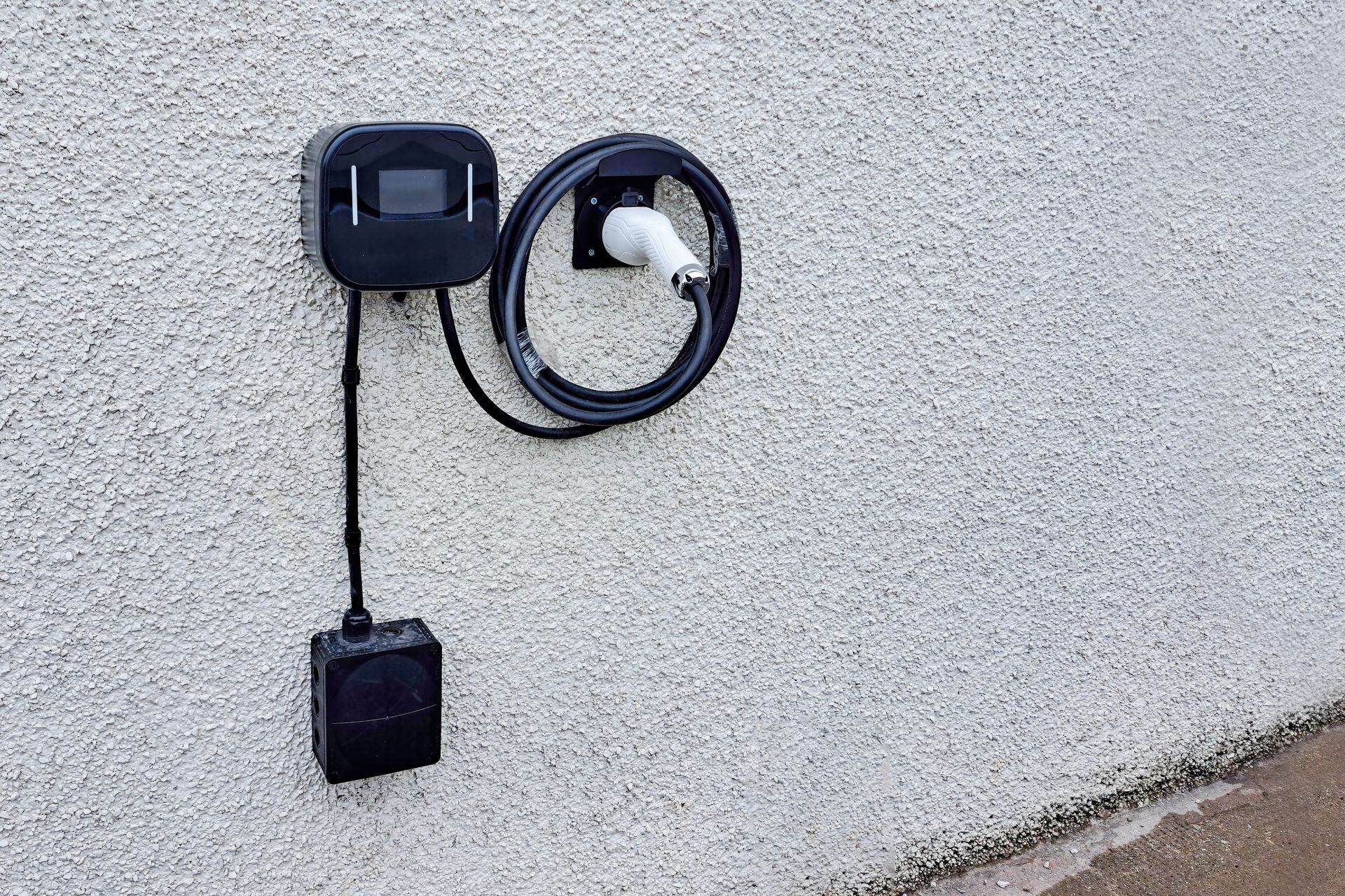 Outdoor wall-mounted electrical outlet and coiled cord on a stucco wall, with a black hanging box.