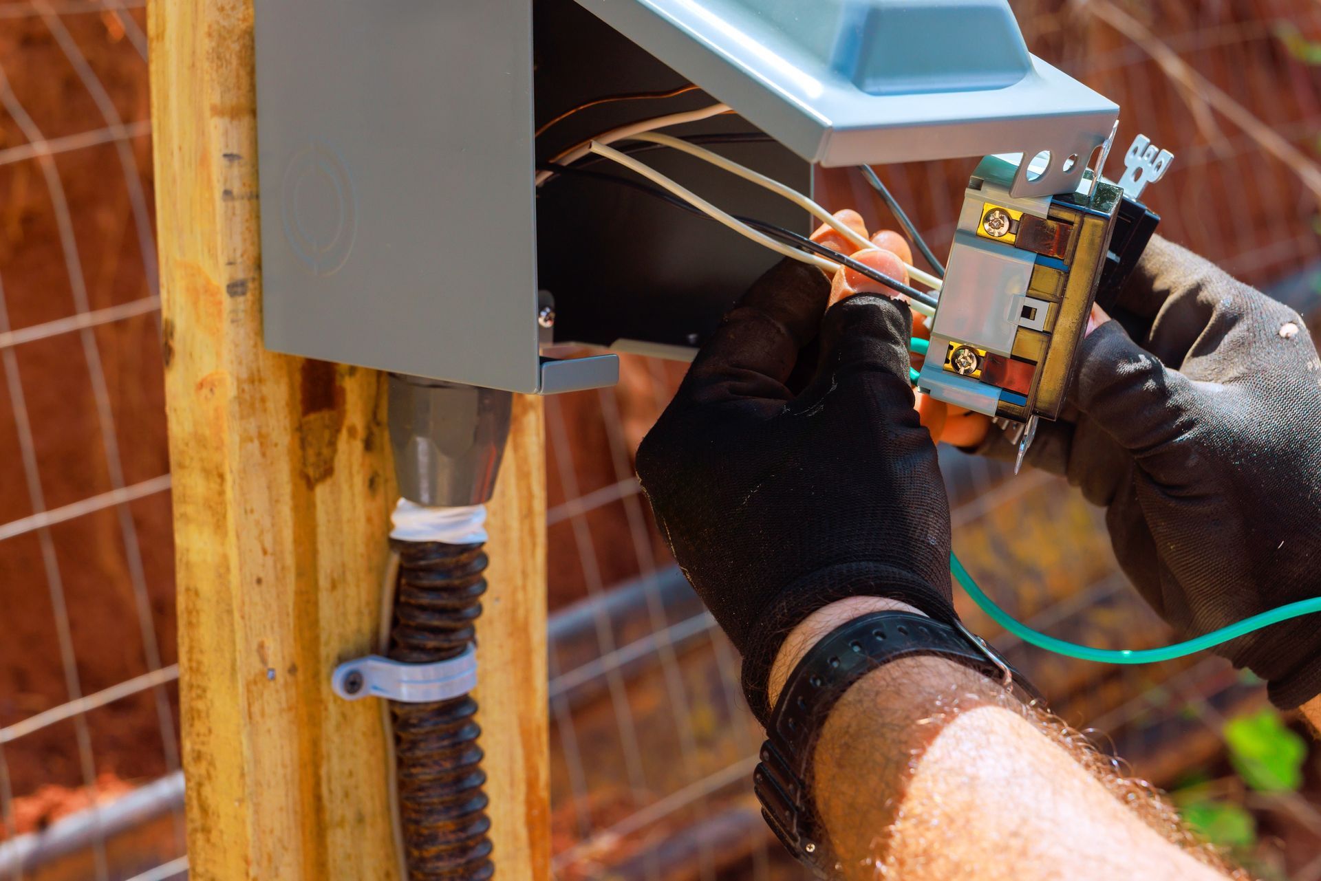 Electrician wiring an outdoor electrical box with gloved hands near a wall-mounted panel.