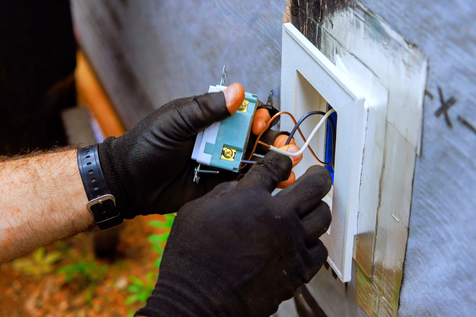 Gloved hands wiring a small electrical device into a wall-mounted panel outdoors