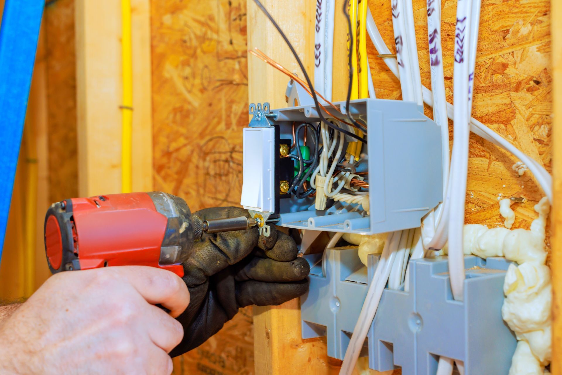 Gloved hand using a drill on an electrical box with exposed wires in a wall cavity