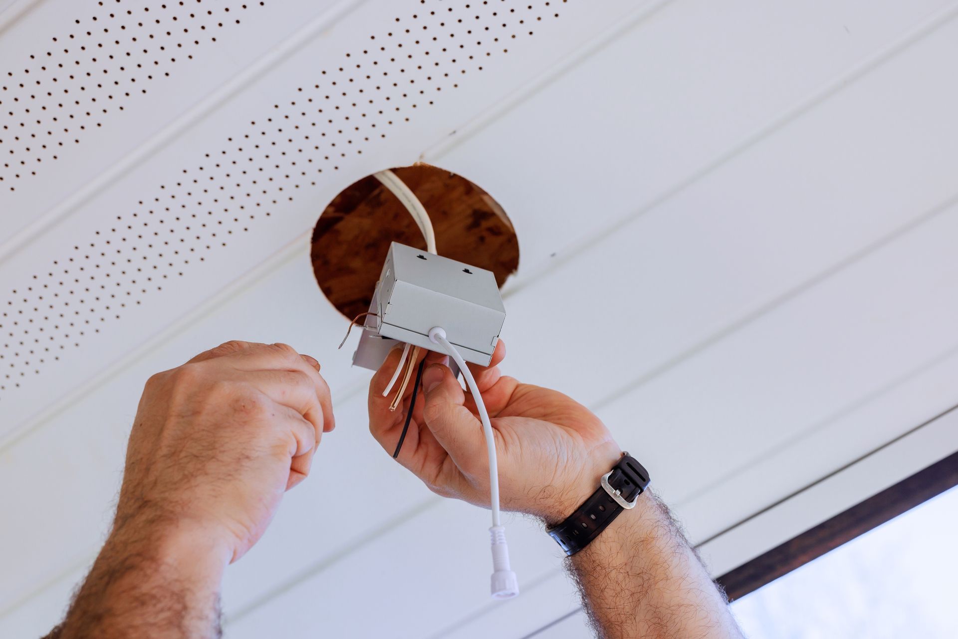Hands installing a white electrical box in a ceiling opening with exposed wires.