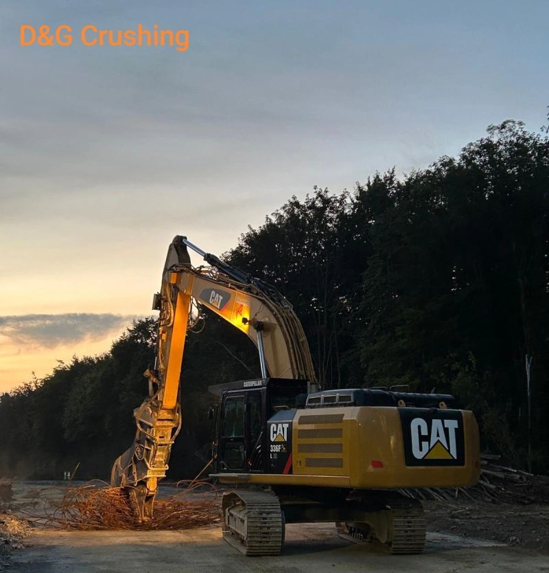 Een CAT-graafmachine verpulvert rotsen in de schemering naast een rij bomen, met oranje werklampen op de arm.