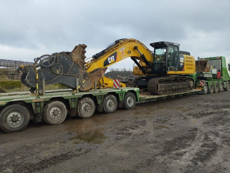 Gele graafmachine geladen op een dieplader op een modderig bouwterrein.