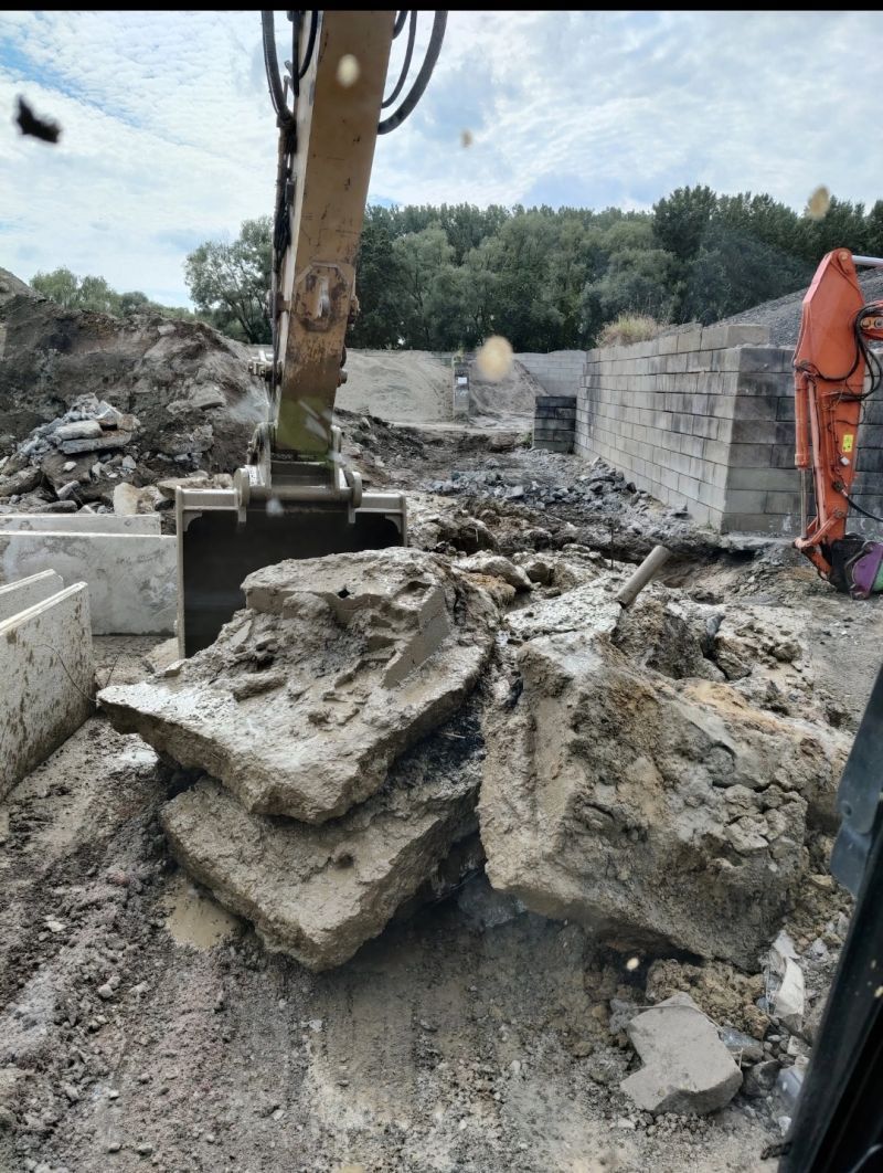 Een graafmachine graaft puin op een sloopterrein, met gebroken beton en ander puin eromheen opgestapeld.