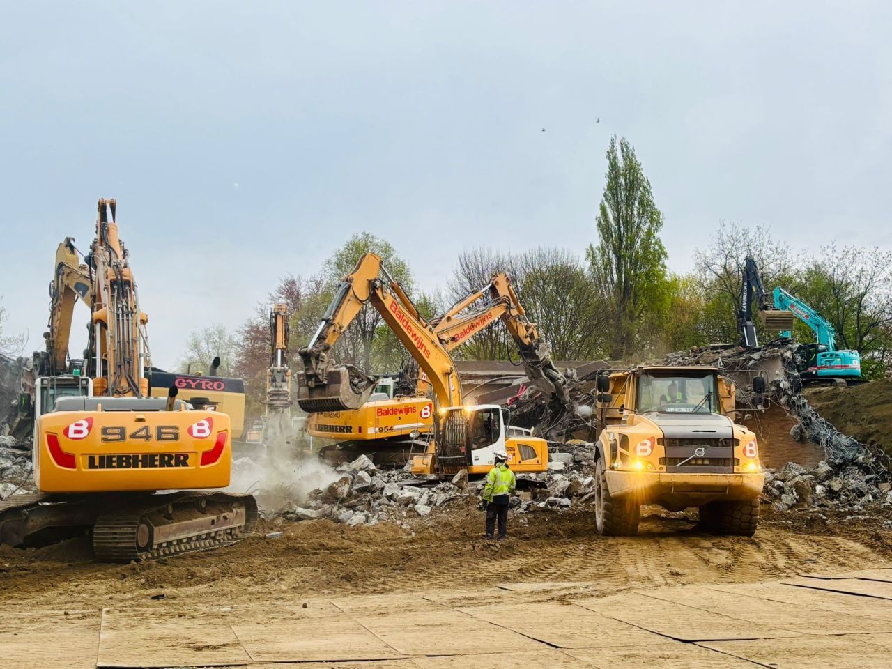 Graafmachines en bouwvoertuigen werken te midden van het puin op een sloopterrein, met stof en een arbeider in de buurt.