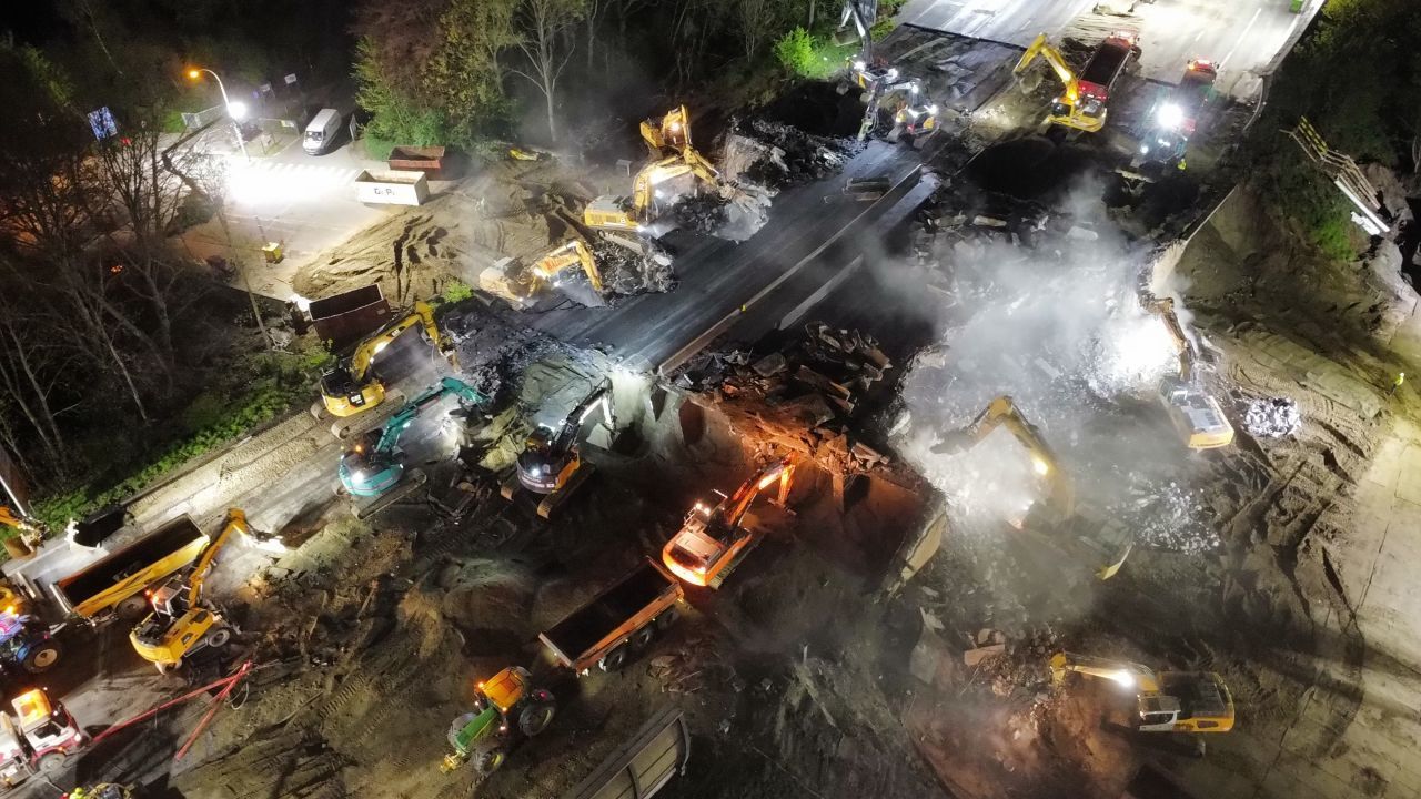 Nachtopname vanuit de lucht van een bouwterrein met graafmachines, vrachtwagens en felle schijnwerpers.