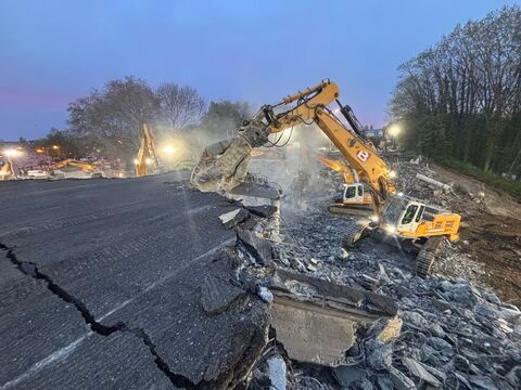 Een graafmachine sloopt in de schemering een stuk gebarsten beton langs de weg, met puin en werklampen.