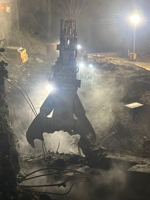 Graafmachinegrijper die puin vastgrijpt op een rokerige bouwplaats 's nachts onder fel licht van de werklampen.