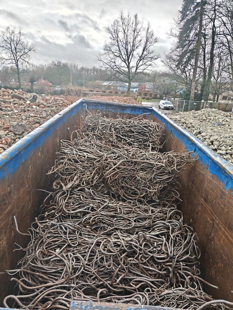 Een blauwe metalen container gevuld met verwarde wortels of takken in een modderige tuin onder een bewolkte hemel.
