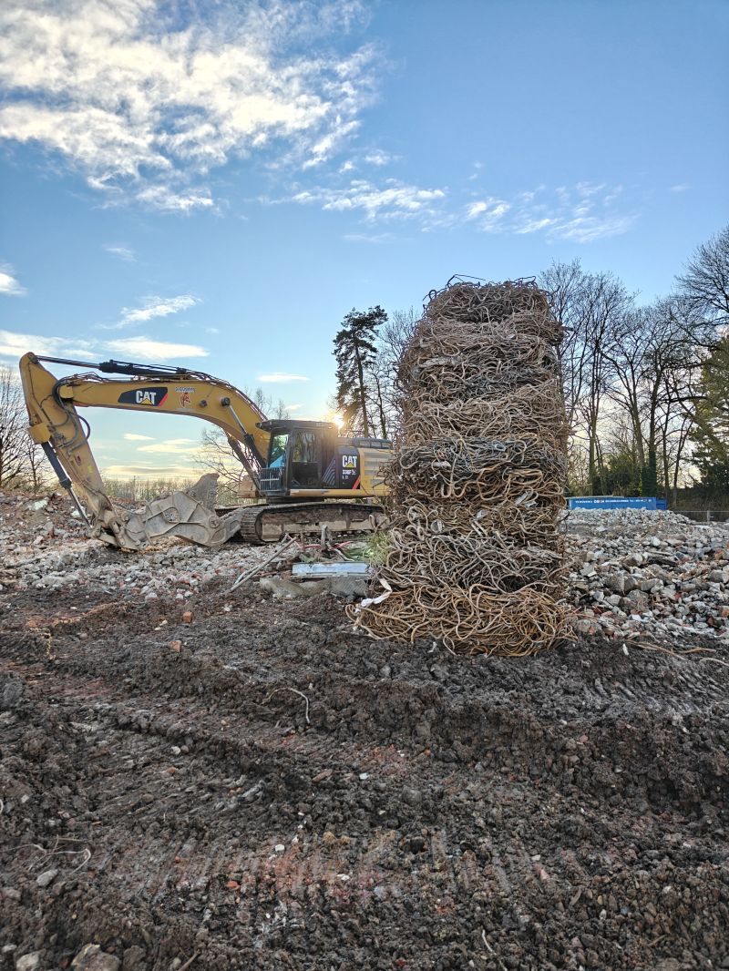 Een graafmachine ruimt een modderig terrein op naast een hoge stapel hout onder een bewolkte hemel.