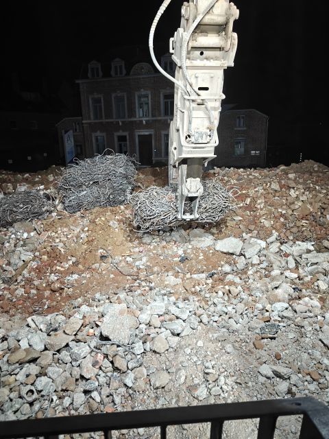 Een boormachine breekt puin op een sloopterrein 's nachts, met een donker gebouw op de achtergrond.