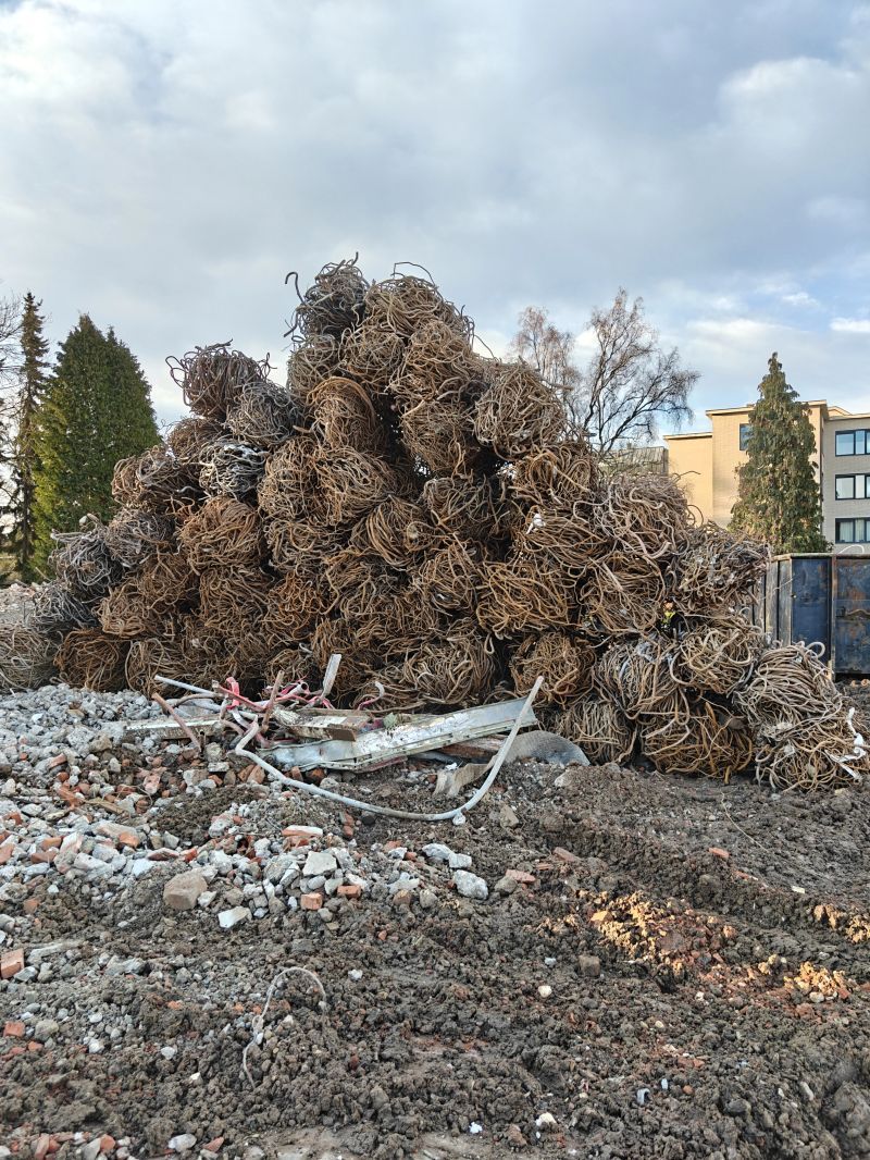 Een grote, ontwortelde boom en een hoop puin liggen opgestapeld op een modderig terrein vlakbij gebouwen.