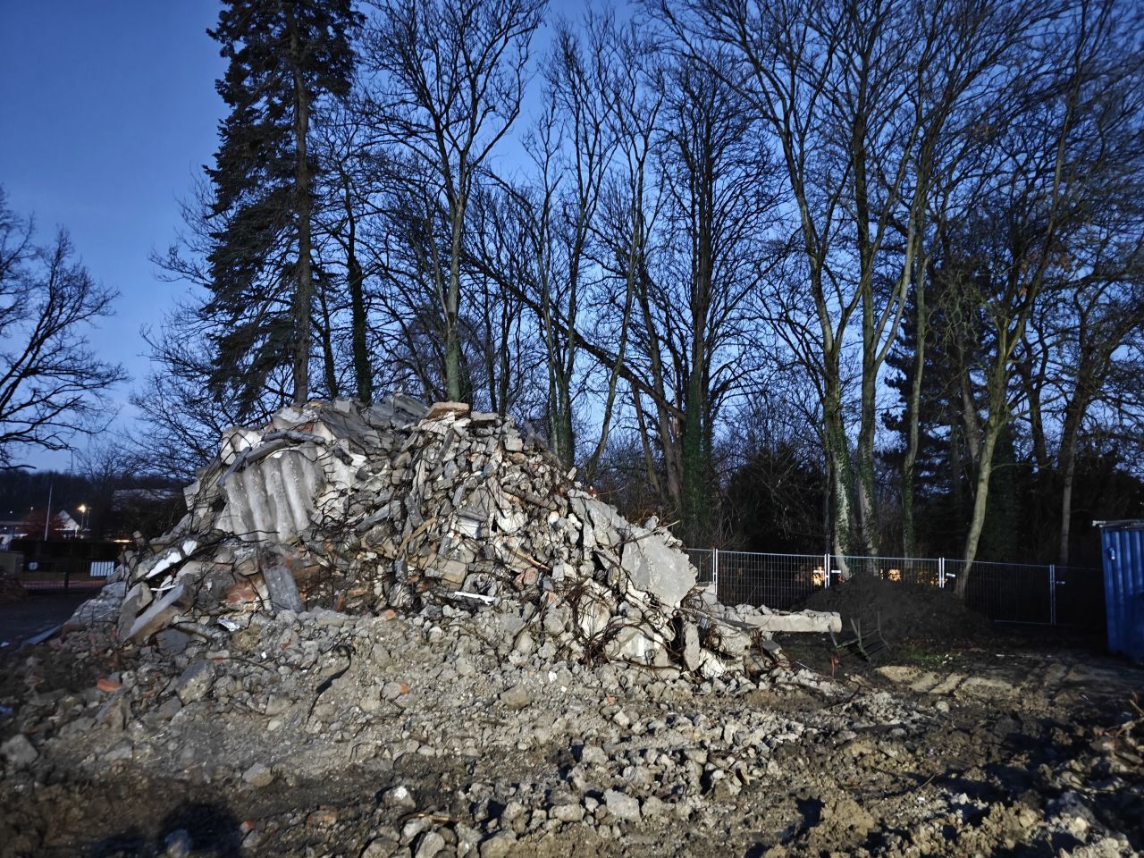 Een grote hoop puin en gebroken beton op een sloopterrein tussen kale bomen in de winter.