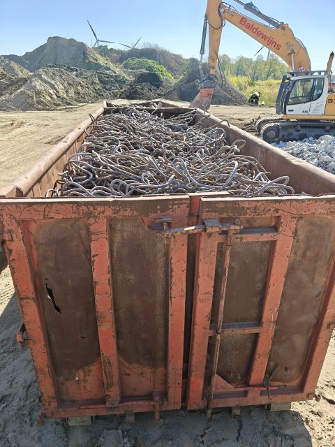 Een roestige afvalcontainer vol wapeningsstaal, met daarachter een graafmachine in een steengroeve.