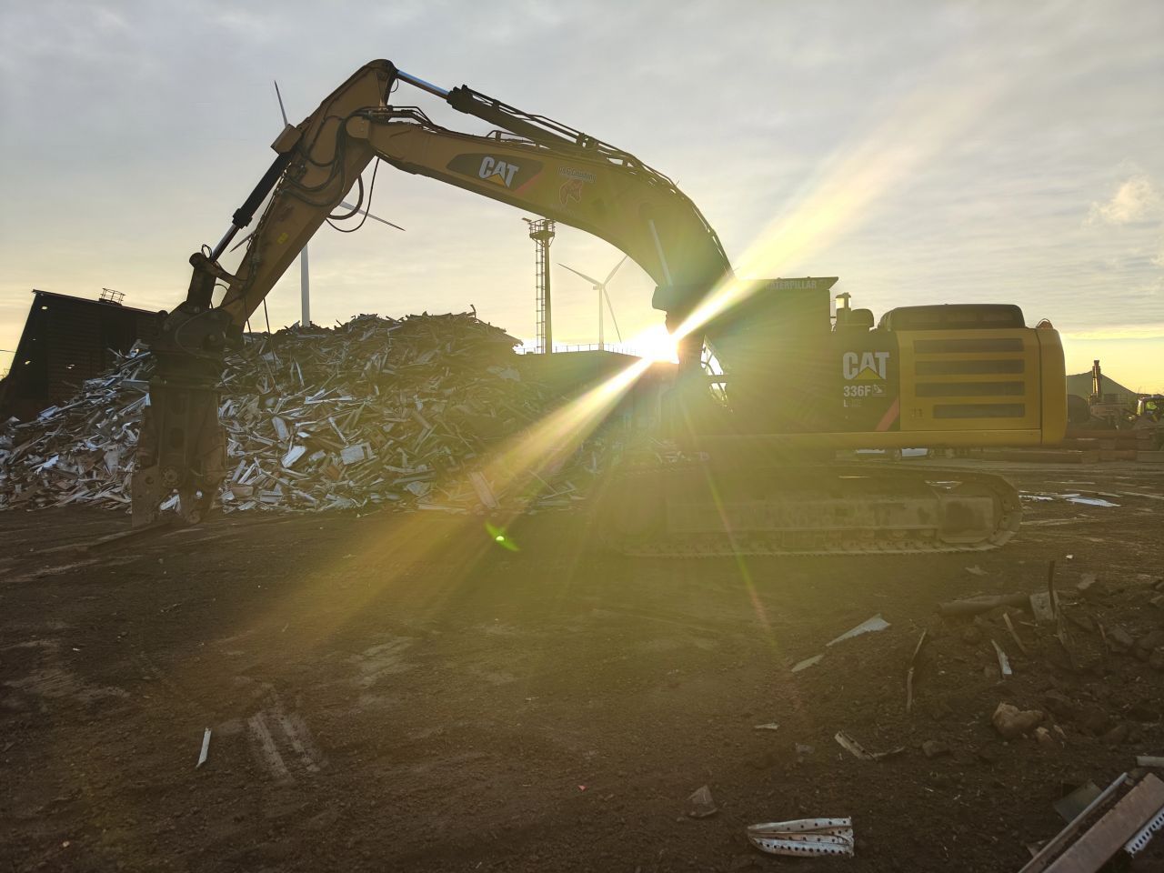 Een graafmachine, afgetekend tegen de zon op een schroothoop, is bezig met het verwijderen van puin bij zonsondergang.