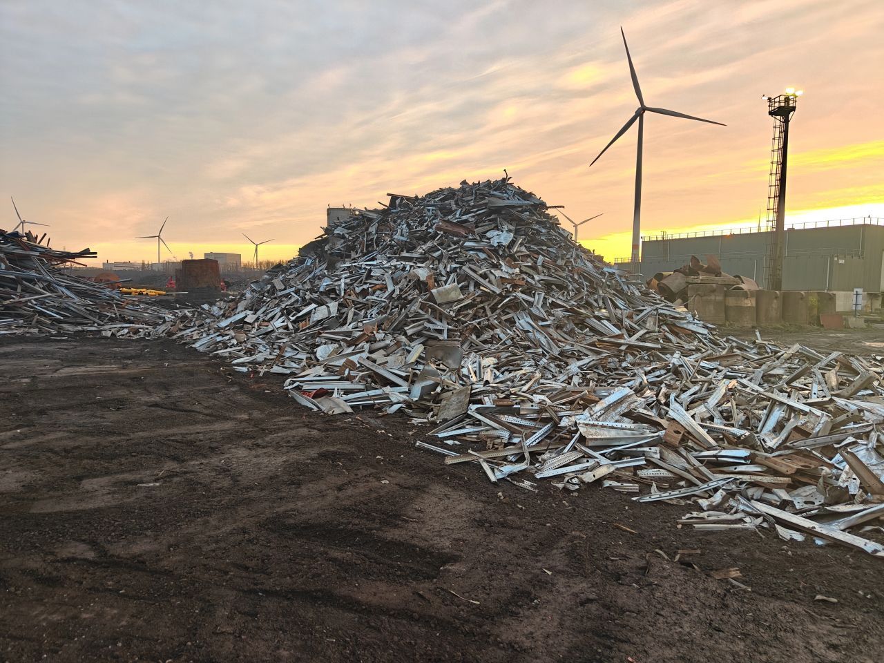 Hopen schrootmetaal op een industrieterrein bij zonsondergang, met een windmolen en een elektriciteitspaal op de achtergrond.