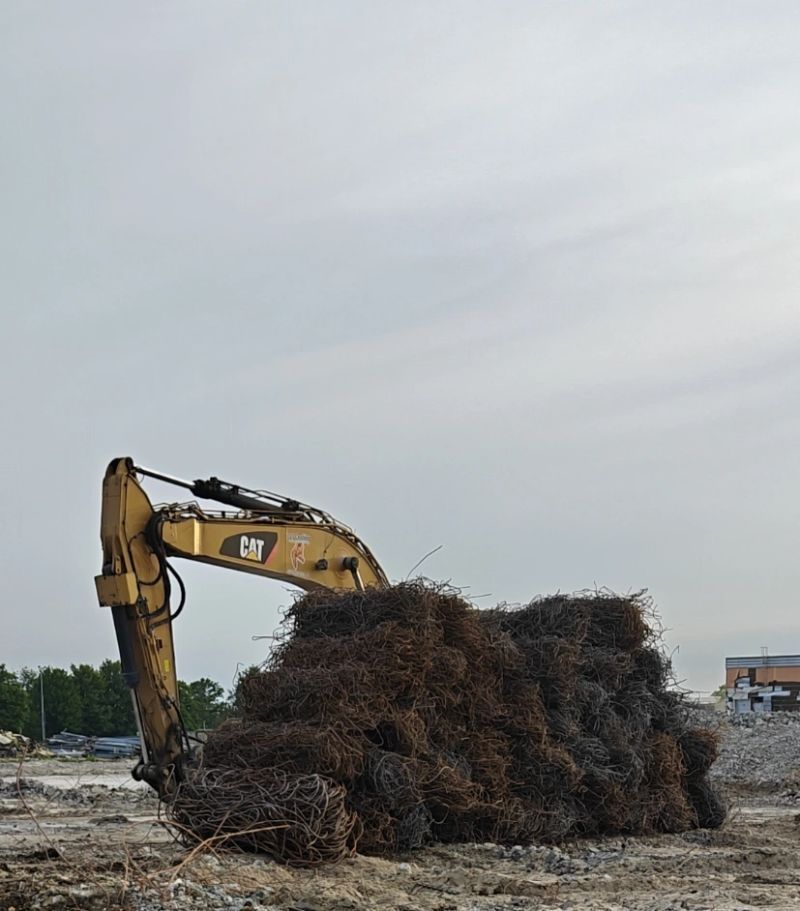 Een gele graafmachine verplettert een grote hoop takken en puin op een bewolkte bouwplaats.
