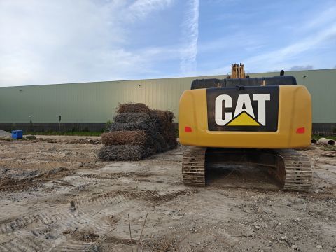 Een gele CAT-graafmachine staat geparkeerd op een onverhard terrein naast een groot groen gebouw met opgestapelde materialen.