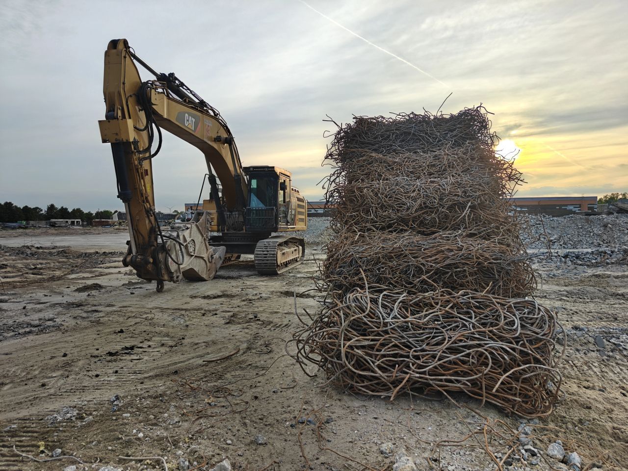 Een graafmachine naast een grote stapel schroot op een bouwterrein bij zonsondergang.