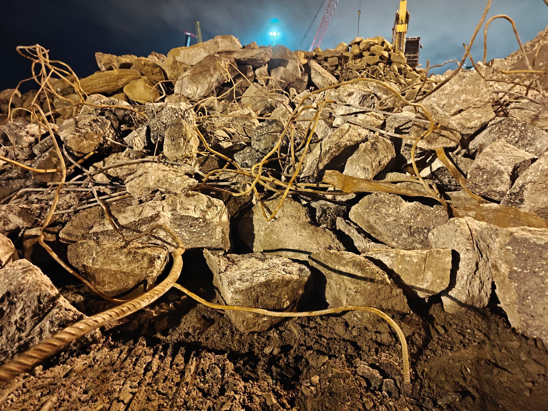 Puin van gebroken beton en verstrengeld wapeningsstaal onder een blauwe hemel, met een kleine kraan op de achtergrond.