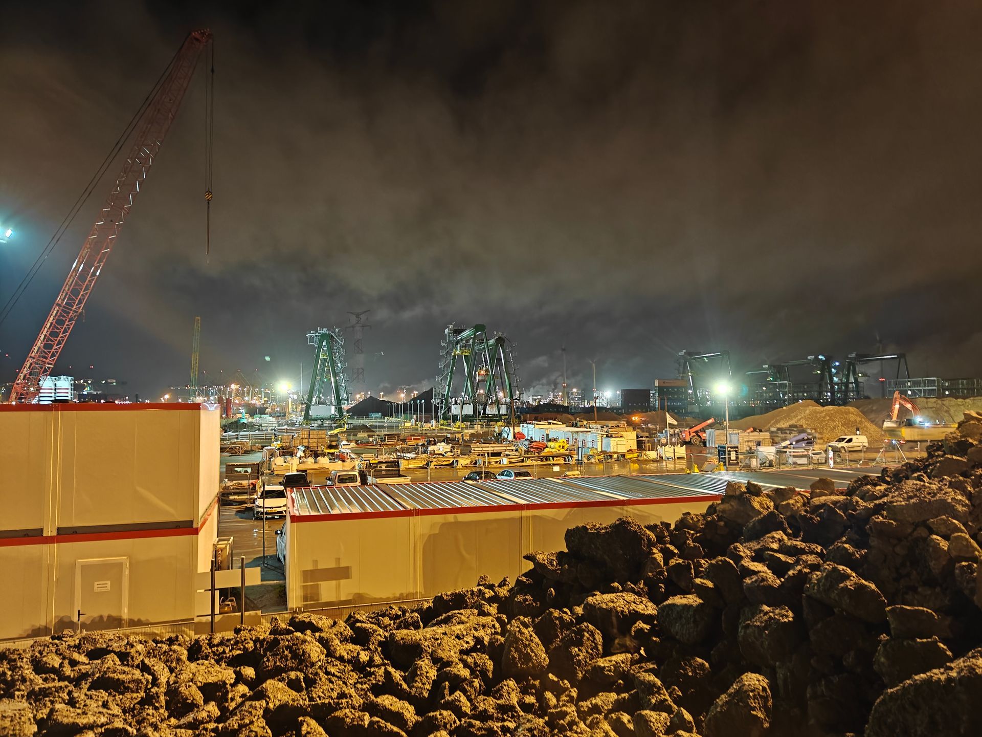 Een bouwplaats 's nachts met verlichte aanhangwagens, zwaar materieel en stadslichten onder een bewolkte hemel.