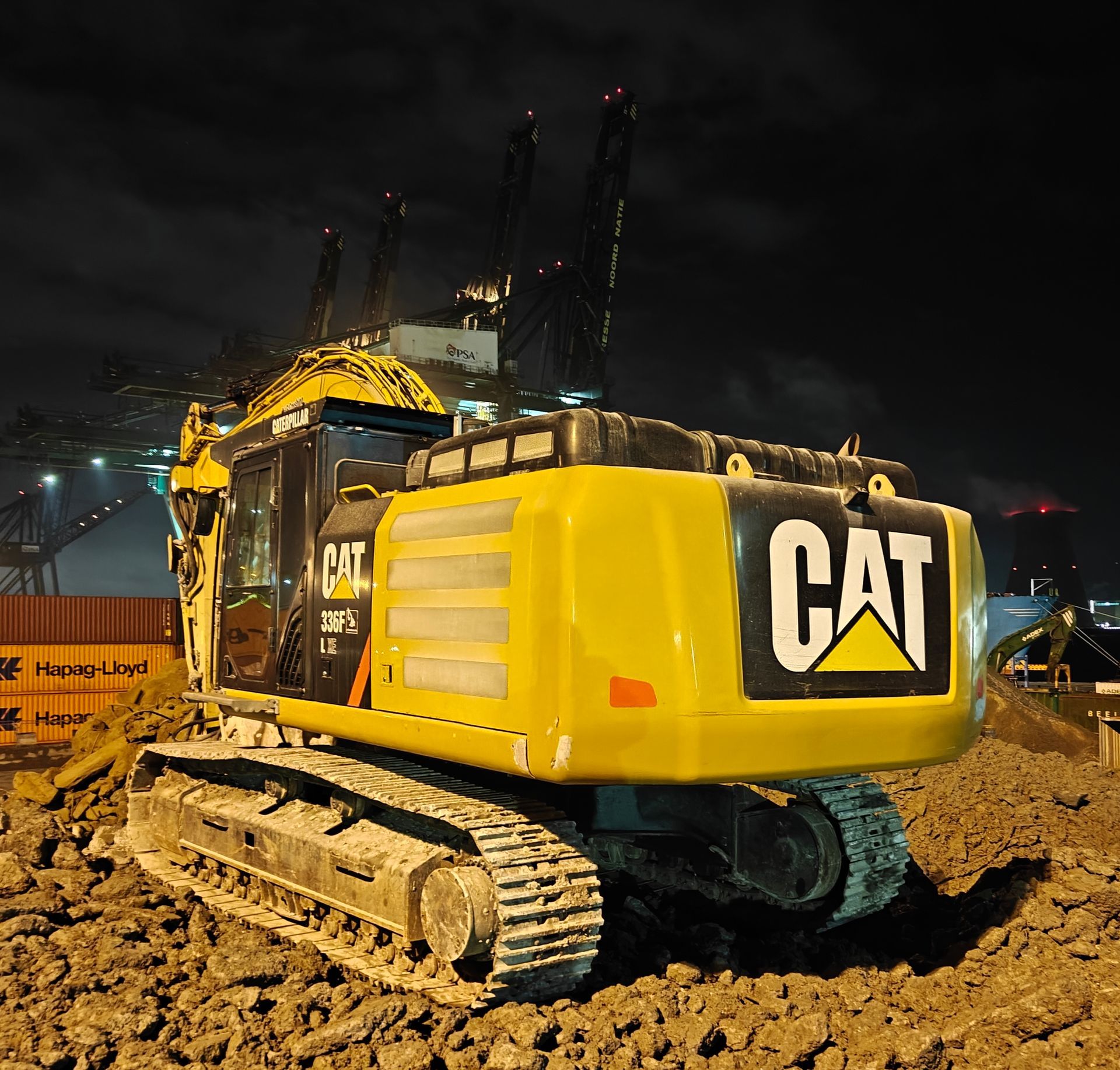 Een gele CAT-graafmachine staat 's nachts op een modderige ondergrond, met bouwkranen op de achtergrond.
