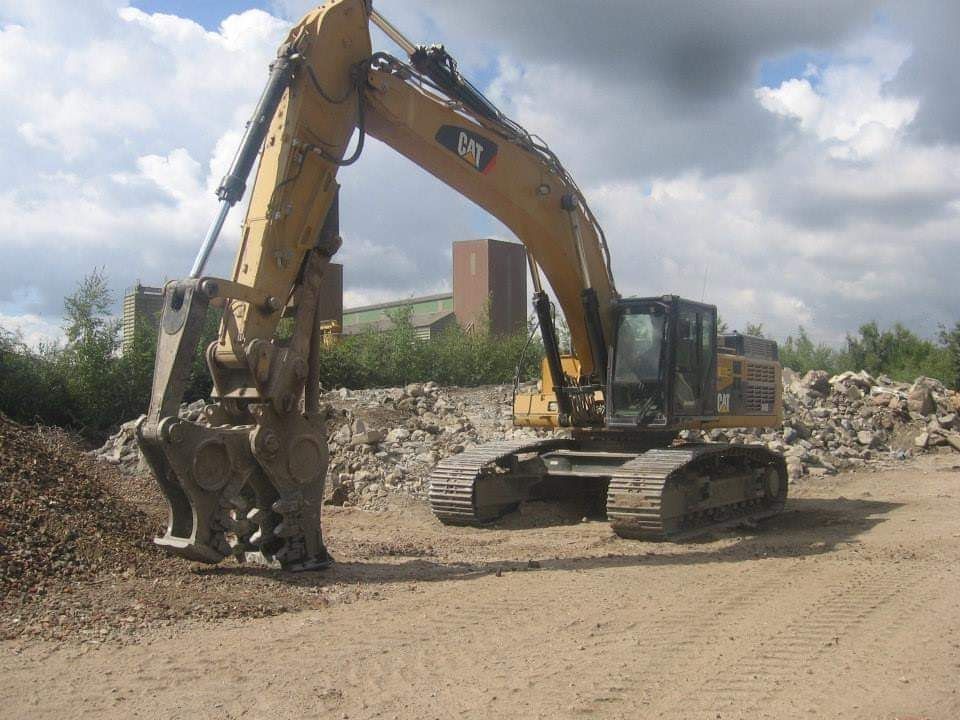 Een gele graafmachine op een onverhard terrein, aan het graven nabij een puinhoop onder een bewolkte hemel.
