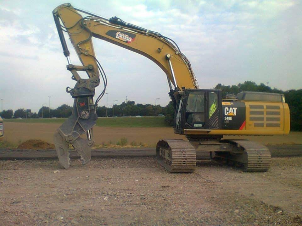 Gele CAT-graafmachine met grijper op een grindterrein onder een bewolkte hemel.