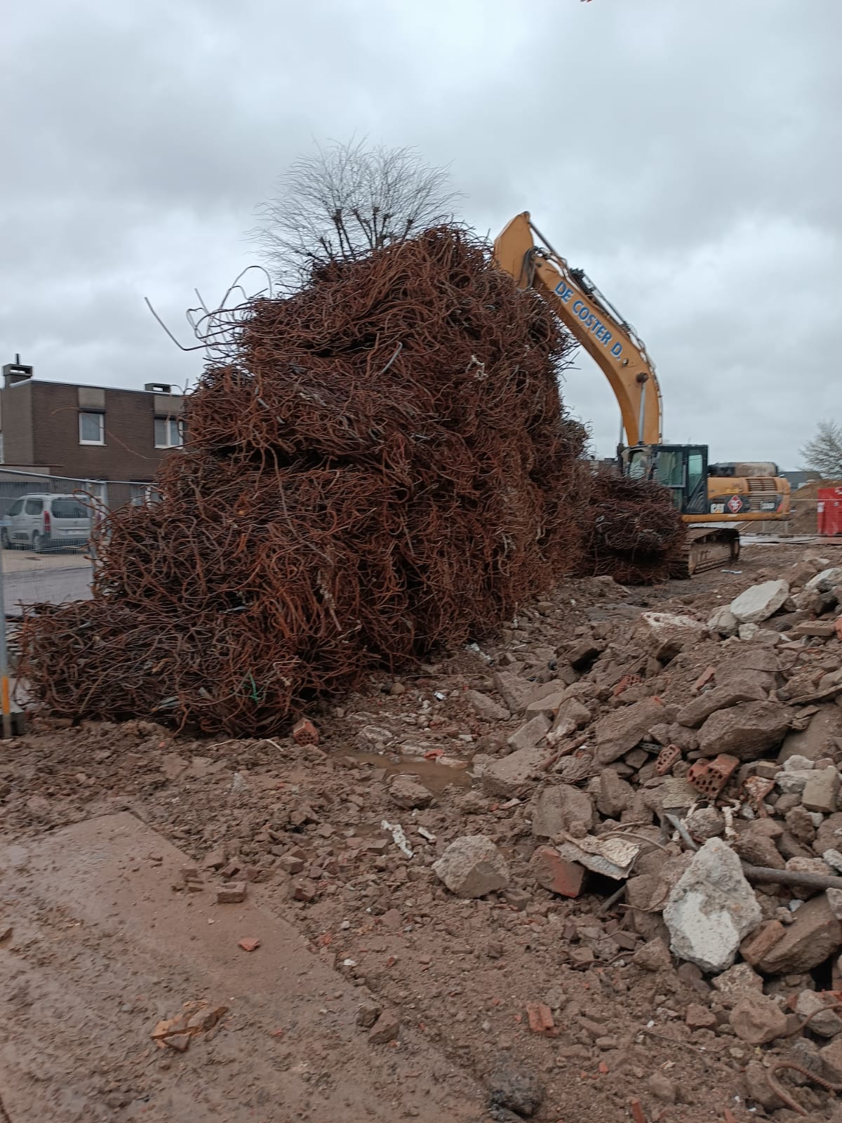 Een graafmachine tilt een grote wortelkluit op van een bouwterrein met puin en gebouwen in de buurt.