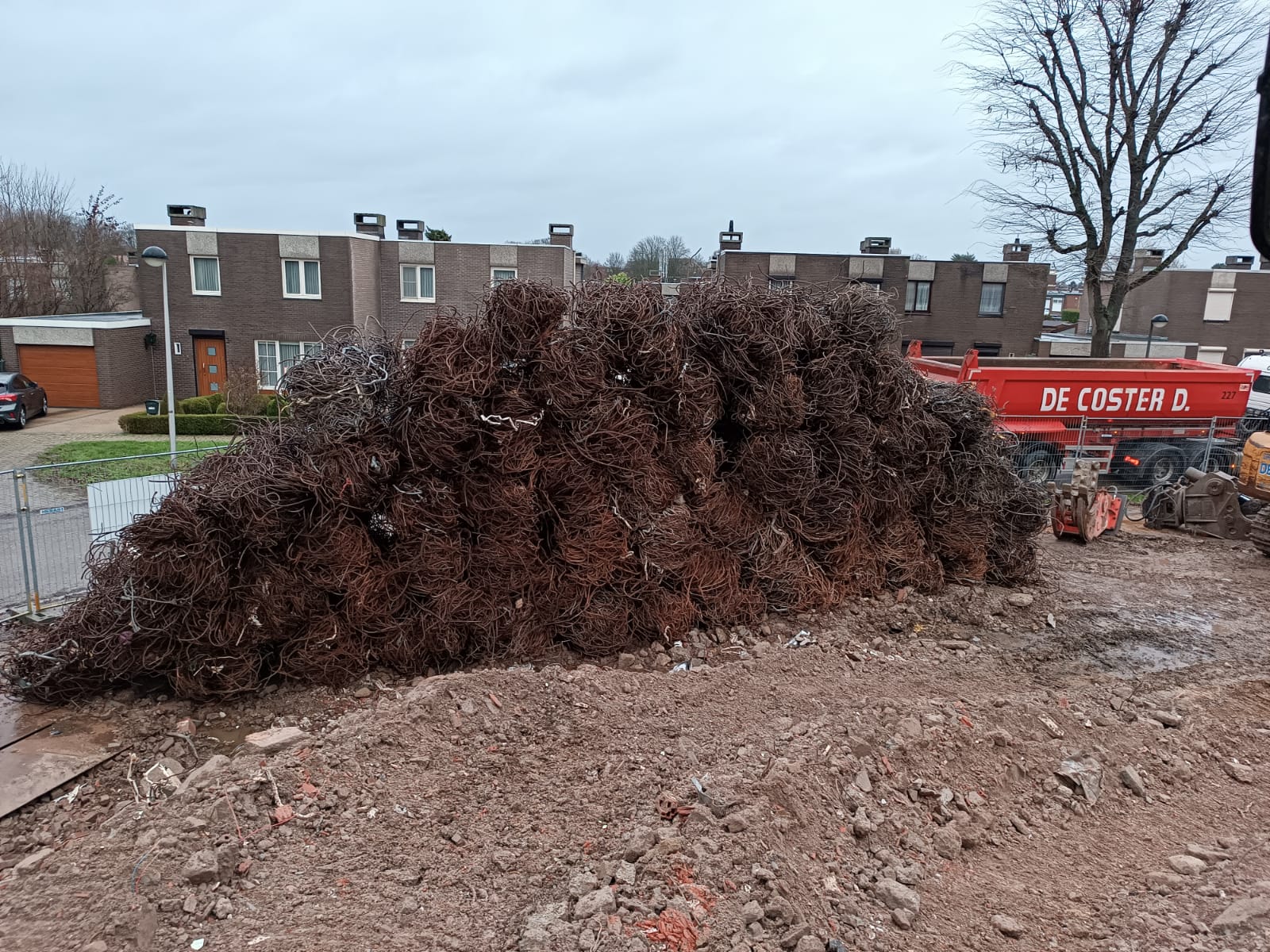 Een grote hoop ontwortelde boomwortels op een modderig bouwterrein met bakstenen huizen in de buurt.