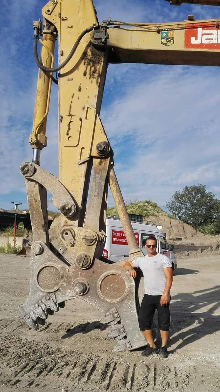 Een persoon poseert naast een grote graafmachineklauw op een bouwterrein, met een modderige ondergrond