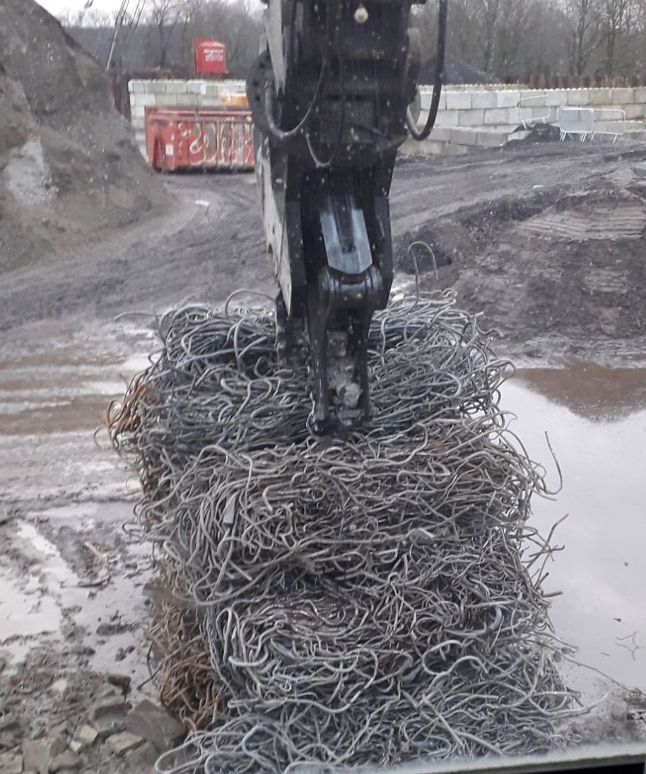 Een graafmachine tilt een verwarde bundel metalen draden op van een modderige bouwplaats.