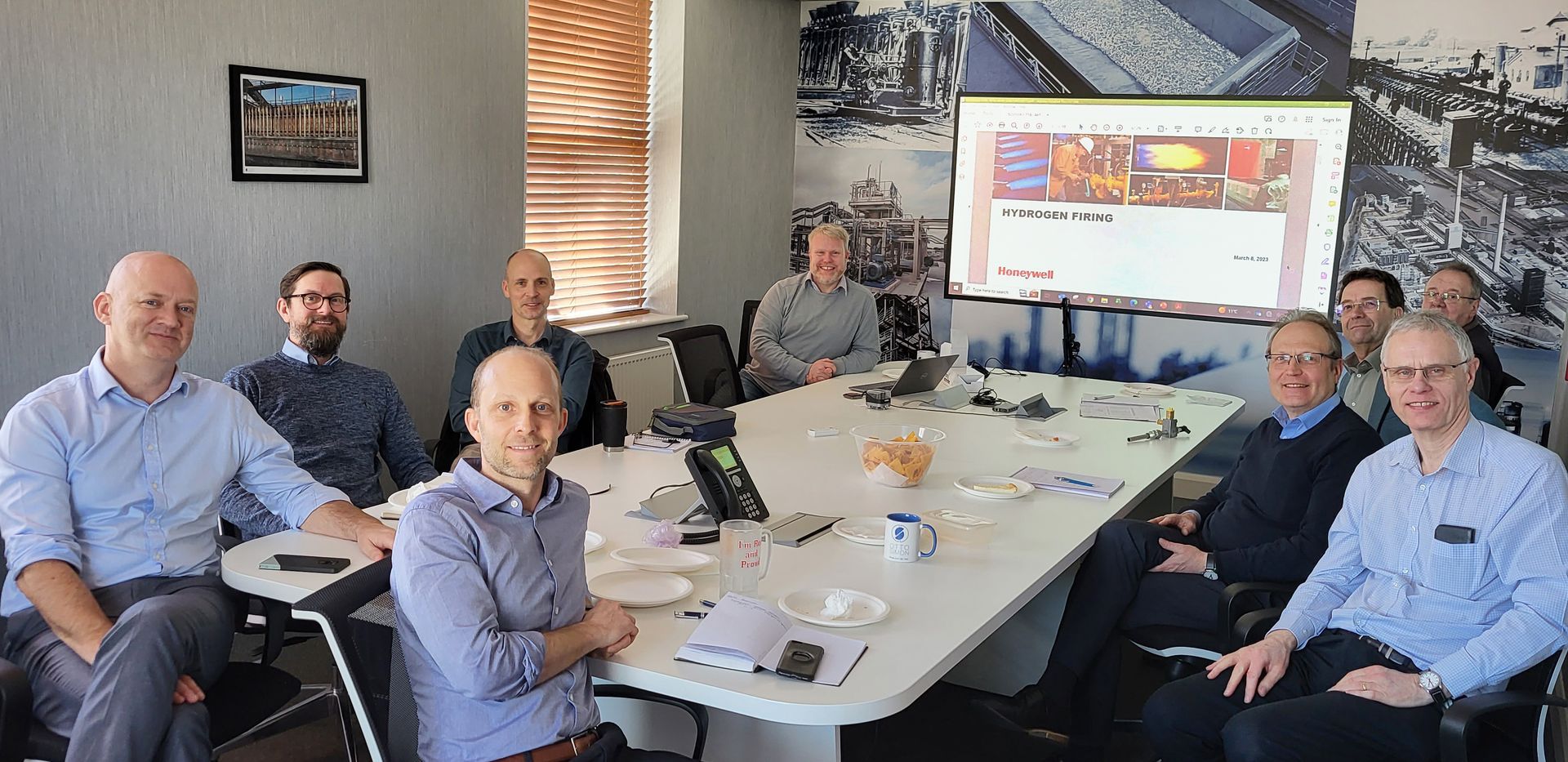 Otto Simon and Honeywell engineers sit around a conference table, turned to face the camera with a 'hydrogen firing' presentation slide projected on the wall behind them.