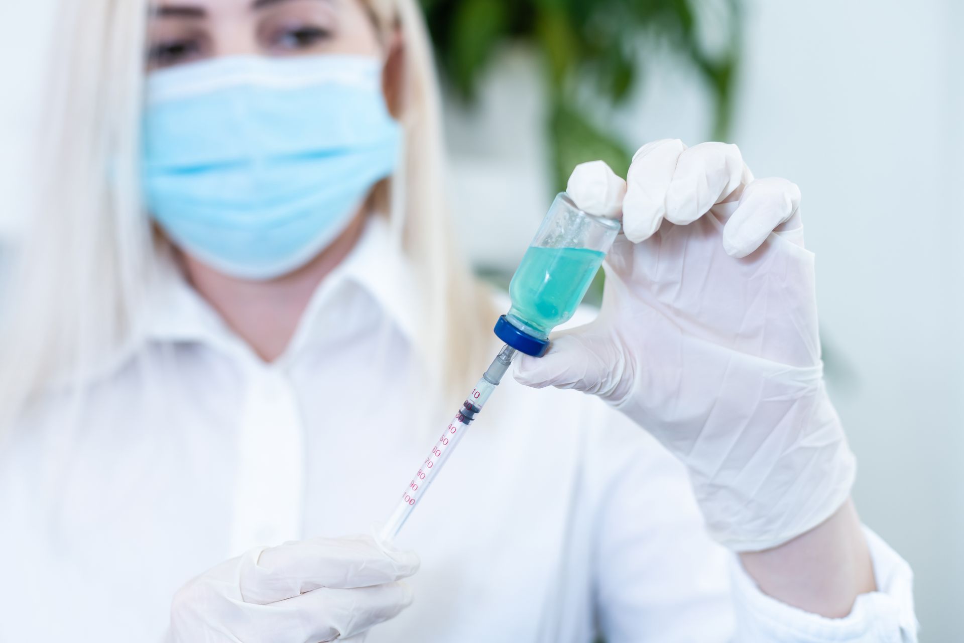 Healthcare worker holding a syringe and vial with blue liquid, wearing a mask and gloves