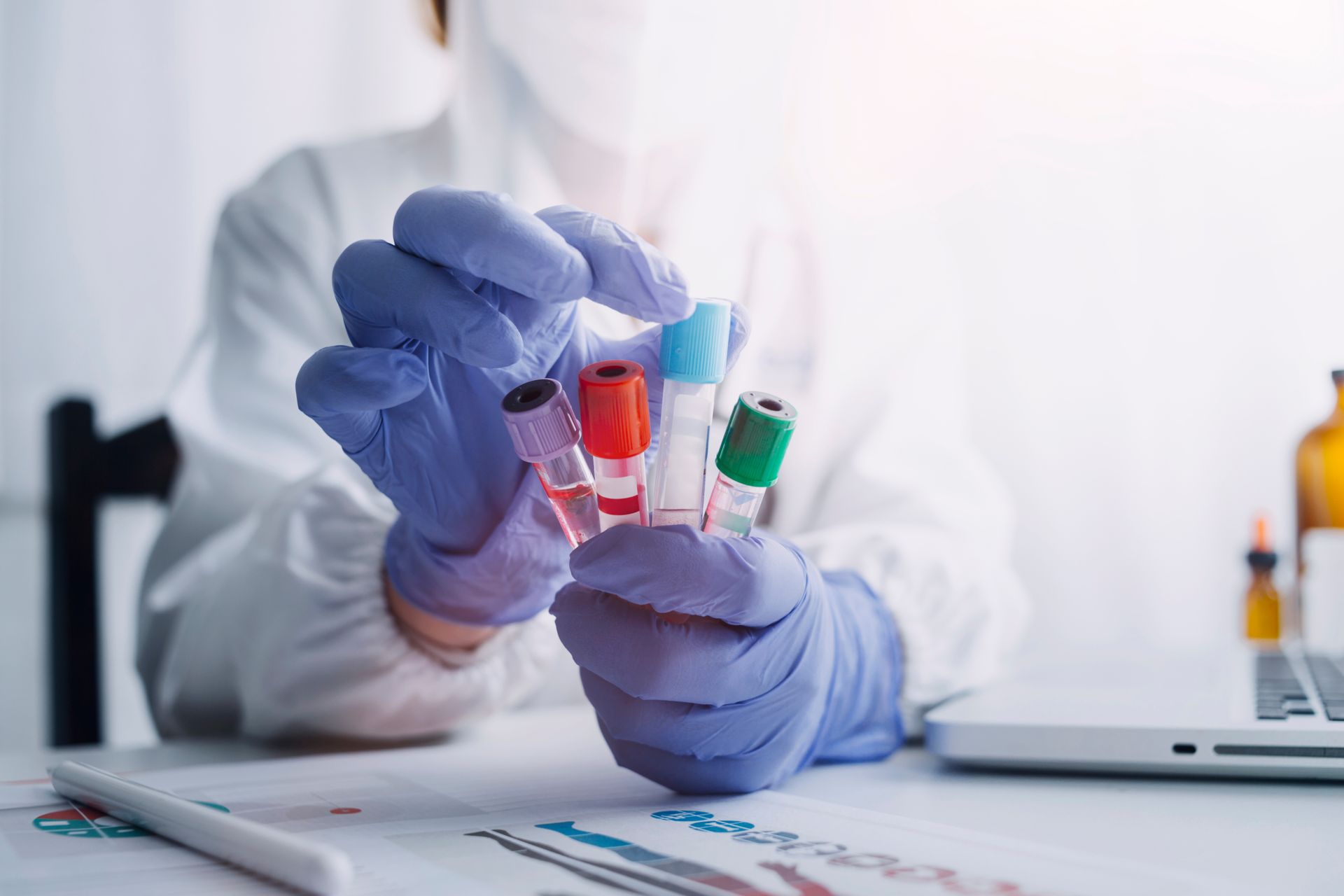 Gloved lab worker holding three colorful test tubes over paperwork in a bright laboratory