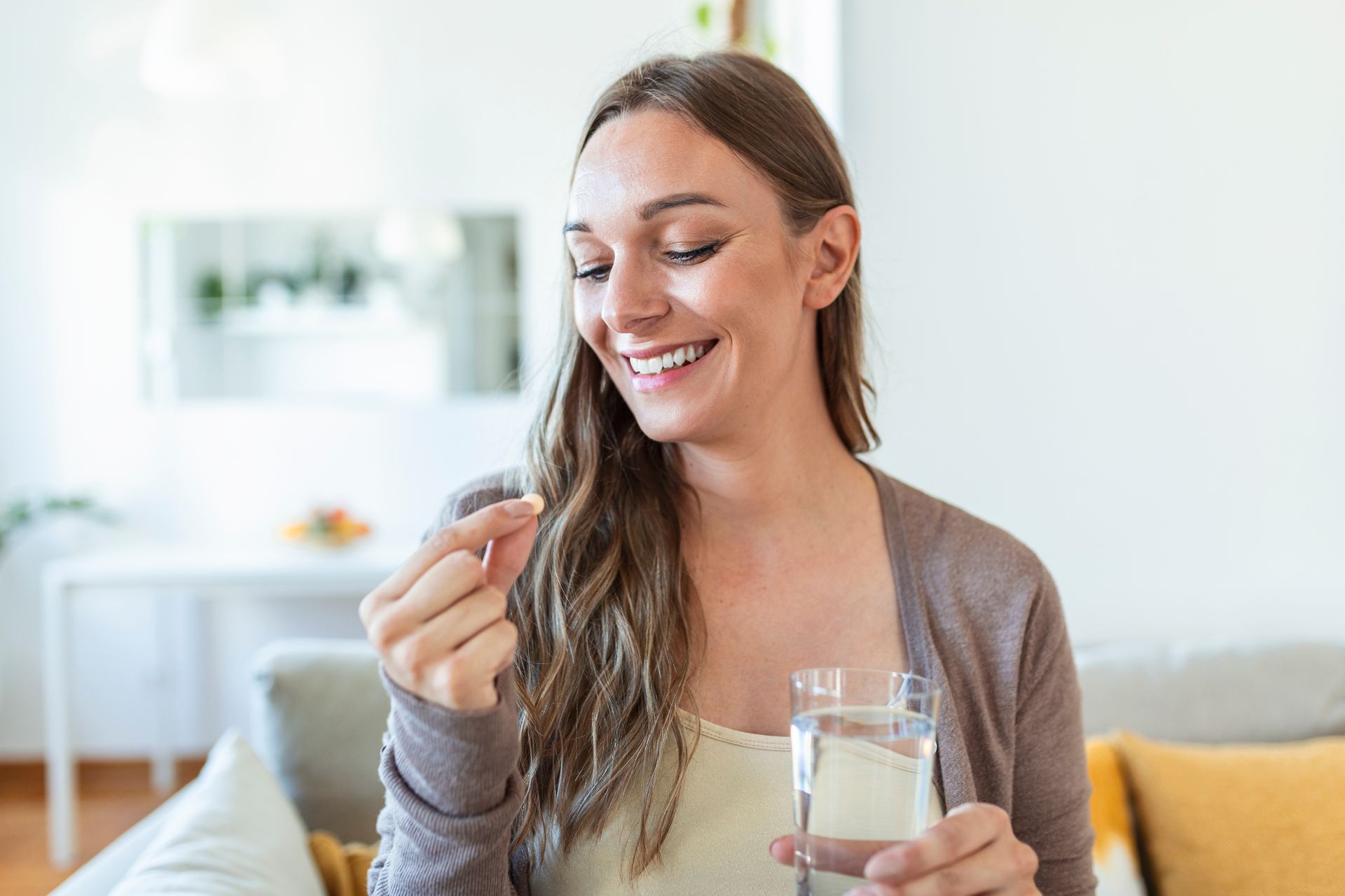 Woman smiling while holding a pill and a glass of water in a bright room