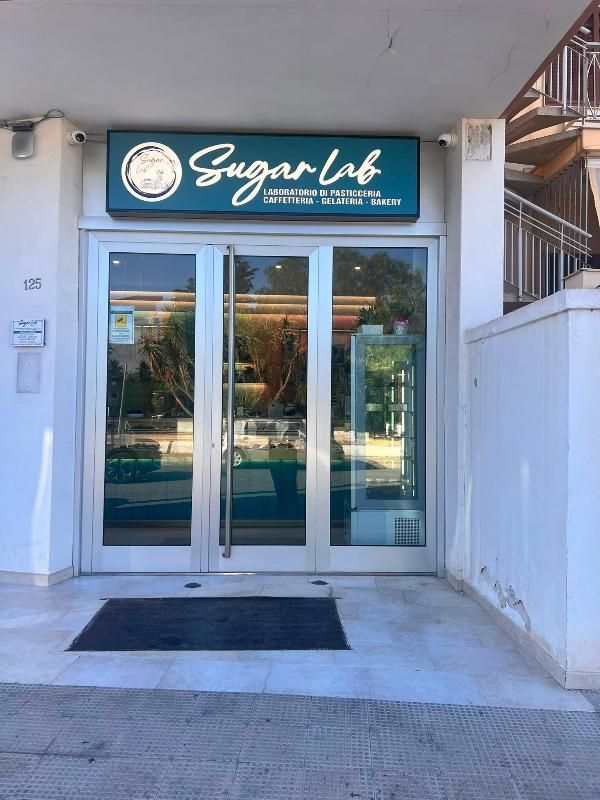Vetrina del negozio Sugar Lab con porte a vetri e insegna color verde acqua su un marciapiede soleggiato